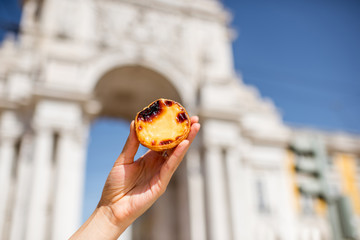 Holding portuguese egg tart pastry called pastel de Nata outdoors on the triumphal arch background in Lisbon