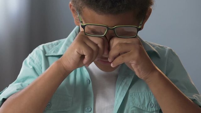 Little biracial boy in glasses doing homework, rubbing eyes, strained eyesight