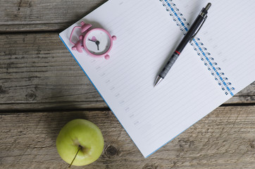 Open notebook agenda with timetable and small pink clock at 8:00 am and green apple and pen on wood