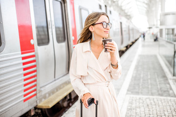 Fototapeta premium Portrait of a young elegant woman standing with coffee cup near the train at the railway station