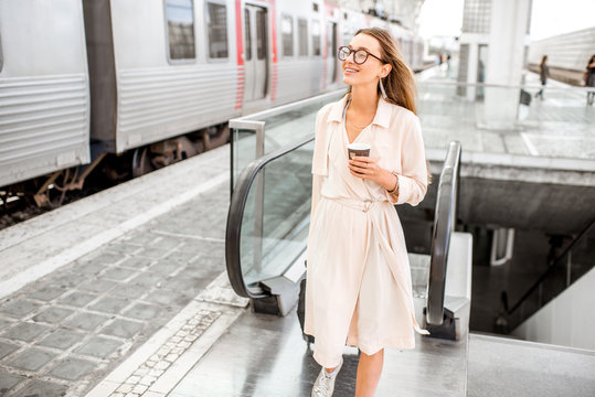 Young and elegant woman walking with luggage and coffee cup near the train at the modern railway station