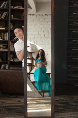 a pregnant young woman in a blue top and a skirt stands near the mirror, a man looks out from behind a mirror and looks at it.Happiness. Expectation. The blue skirt.
