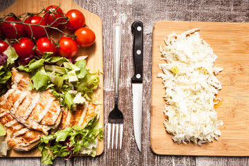 Grilled chicken breast on fresh cut salad next to a bunch of tomatoes on a wooden board. On the second wooden board is cabbage salad
