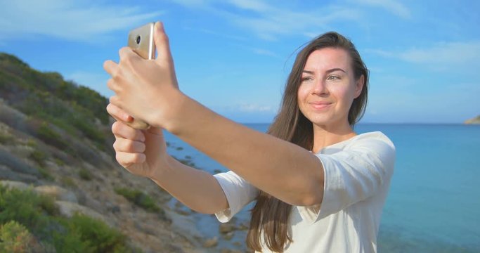 Young pretty woman doing selfie photo using her smartphone over sea. Female standing near the sea during sunset. Clear blue water.