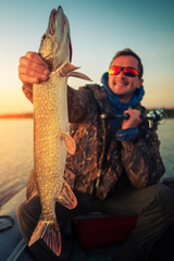 Naklejka premium Happy angler holds the pike fish with natural sunrise lake background