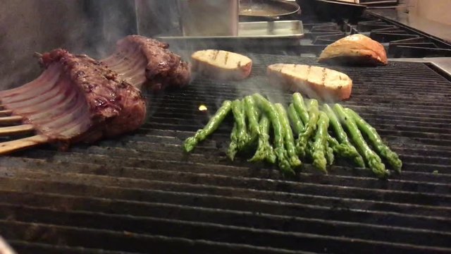 Asparagus, lamb and bread line a grill where a chef uses tongs thereafter to move around vegetables with smoke and steam emitting upward.  