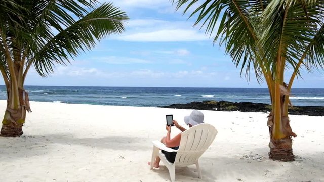 Woman relaxing on vacation sitting on tropical beach under palm trees on deckchair reading an electronic book or e-reader. Filmed in Samoa, a South Pacific island