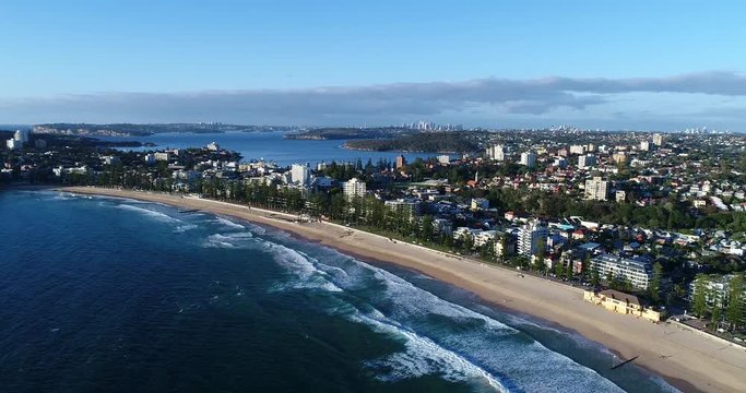 Manly beach waterfront backwards flying in view of promenade, streets, trees and Sydney CBD in background.
