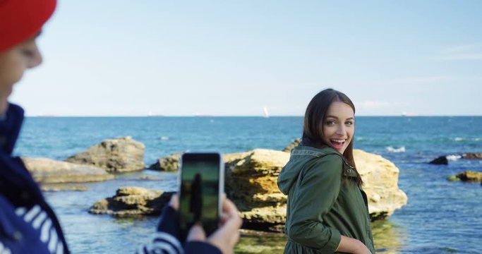 Woman taking nice photos on her mobile phone of her attractive female friend smiling and giving thumbs up on the rocks and sea background. Cold but sunny weather. Outdoors