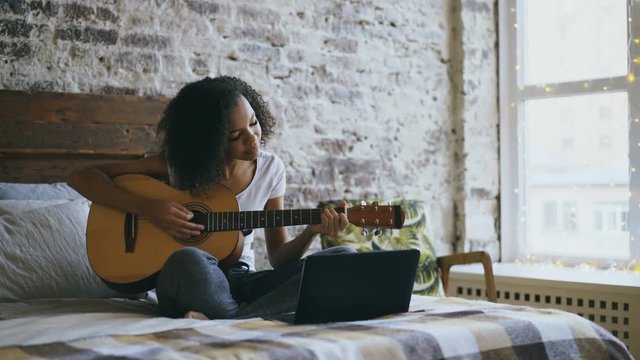 Curly african american teenager girl concentraing learning to play guitar using laptop computer sitting on bed at home