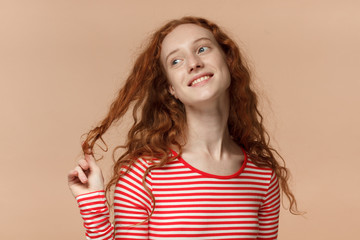 Indoor closeup of young redhead European female pictured isolated on peach background dressed in striped T-shirt twisting her lock in fingers, looking upwards and aside while dreaming and smiling