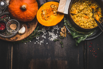 Cooking pot of vegetarian pumpkin risotto and spoon on dark rustic kitchen table background with cooking ingredients, top view, border. Healthy clean seasonal food and eating concept