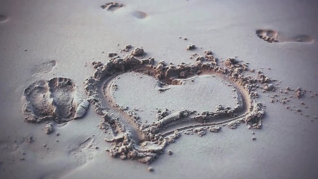 Drawn heart shape in the sand and walking female feet and waves clearing feet traces. 1920x1080