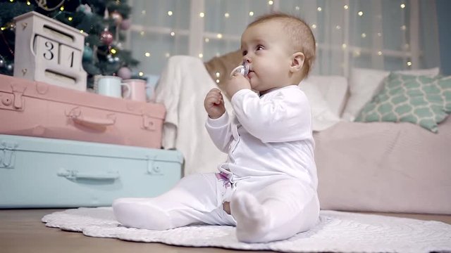 cute infant baby girl in a white jumpsuit sitting on the floor on the Christmas tree. itchy gums will soon be teeth trying to chew on a pacifier