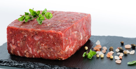 Closeup view on piece of raw ground beef on a black slate board with whole pepper pink salt and fresh marjoram leaves