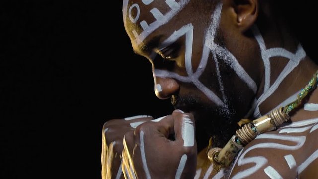 A black man with inflated chest in white patterns gets into a boxing pose and looks into the camera, slow motion