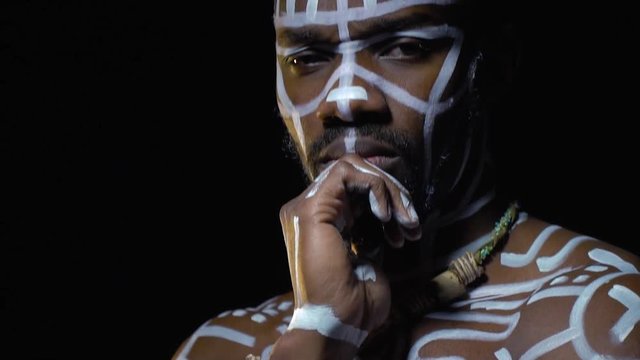 A thoughtful black man with a beard and white ritual patterns brings his hand to his chin and looks into the camera, bit slow motion