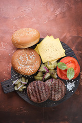 Grilled beef burger cutlets, white sesame buns, pickles with red onion, cheese and tomatoes on a black wooden cutting board. Flat-lay on a rusty metal surface