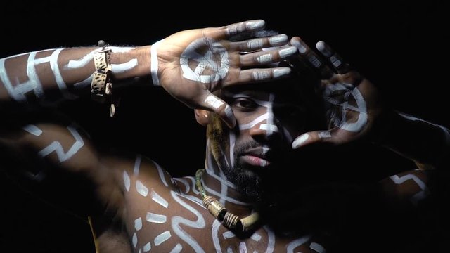Black man in white patterns on the body covers his face with his hands standing in the dark, slow motion
