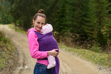 A tiny baby, sleeping peacefully on her mother in a baby sling, and a young beautiful mother walks in nature.