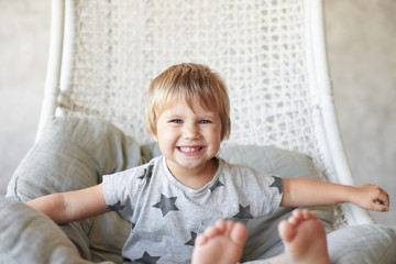 Fototapeta premium Relaxed carefree little girl with short haircut sitting on woven armchair wearing grey t-shirt with stars and no socks, grinning exaggeratedly, showing straight teeth at camera. Selective focus