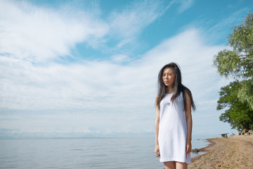 Serious beautiful young Japanese female wearing white summer dress, standing on sandy beach by vast ocean, having pensive look, thinking about her life and future or trying to find solution to problem