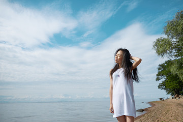 Ecstatic young Asian brunette woman enjoying vacations by the sea, closing eyes and smiling joyfully, feeling happy and carefree standing against blue sky background with copy space for your content