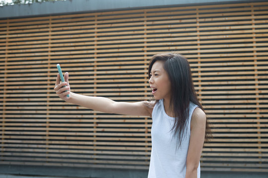Picture of cheerful cute Vietnamese girl having video conference call with friend, using 3g or 4g internet connection on her mobile phone, opening mouth in excitement, extending arm with gadget in it