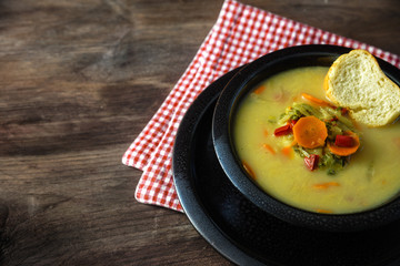 Vegetable cream soup with carrot, red pepper and leek in a black speckled bowl on a red white napkin, brown rustic wooden table with large copy space