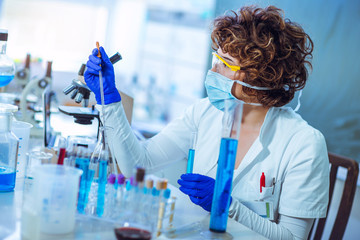 Young female scientist in protective glasses and gloves dropping a liquid substance into the test...