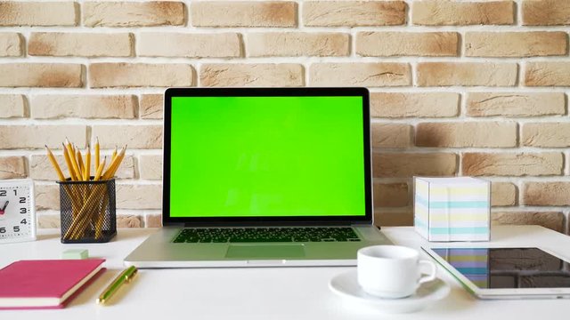 Workplace of a businessman. Laptop with green screen on a table, tablet pc, smartphone, notebook and other accessories on brick wall background. 4K video with close view and very smooth dolly shot.