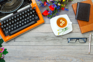Workspace with vintage orange typewriter. Bouquet of summer field flowers, glasses, notebook and cup of chamomile tea.