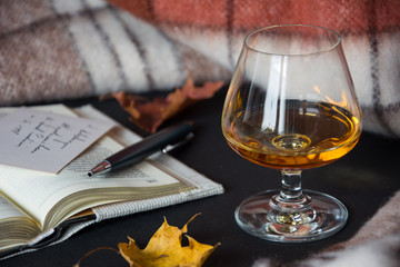 A glass of amber alcohol with an open book, dry leaves, a ballpen and warm blanket in background