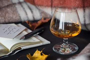 Photo sur Aluminium Alcool A glass of amber alcohol with an open book, dry leaves, a ballpen and warm blanket in background
