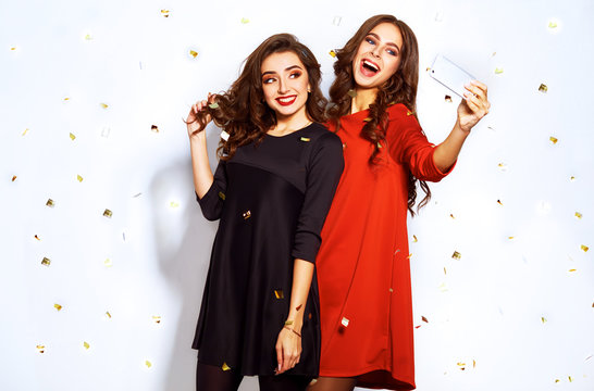 Portrait Of Two Happy Young Women Who Make Selfie. In Evening Dresses On Party Over White Background. Firecrackers In The Background.confetti.the Concept Party.funny Faces
