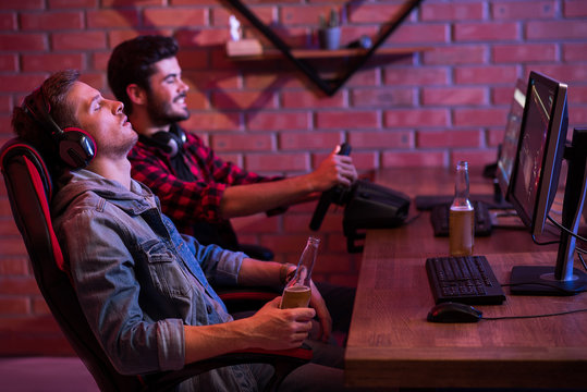 Profile of young gamers are sitting at desk. Focus on sleeping guy is resting in chair with bottle of beer. Cheerful bearded man is enjoying car simulator with steering wheel in background. Copy space