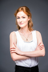 portrait of a young redhaired beautiful girl in the studio on a gray isolated background. A woman is standing with her arms folded and smiling in a white shirt with a short sleeve. Business concept