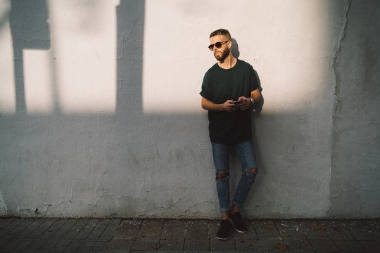 Handsome bearded guy holding a smartphone while standing on a white wall background on a street. Stylish man wearing sunglasses surfing the web while waitinig for a friend to walk the city.