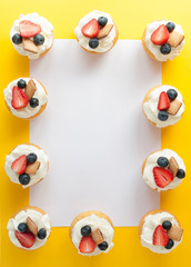 Obraz premium Cupcakes with cream, strawberry, blueberry and cookie on yellow background in the form of a frame. Image with copy space for the confectionery menu, cards, greetings, birthday invitations. Top view