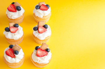 Cupcakes with whipped cream, strawberry, blueberry and cookie on yellow background arranged in ranks. Image with copy space for the confectionery menu, cards, greetings, birthday invitations. Top view