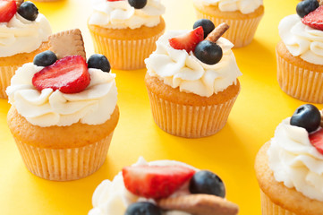 Cupcakes with whipped cream, fresh strawberry, blueberry and cookie on yellow background arranged in ranks. Image for the confectionery menu, cards, greetings, birthday invitations.
