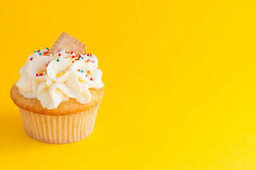 Cupcake with whipped cream, colorfull confectionery sprinkles and cookie on yellow background. The image with copy space. Background for the confectionery menu, cards, greetings, birthday invitations.