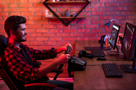 Profile of bearded young positive man is sitting at table and playing car racing video game and steering wheel. He is expressing excitement and gladness. Brick wall in background. Copy space