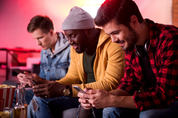 Cheerful news. Selective focus of joyful young man is sitting on sofa with mobile phone while looking at screen of gadget and expressing gladness. His friends are holding smartphone in background