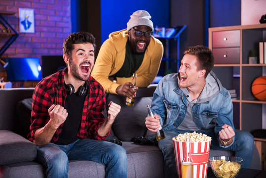 Goal. Delightful friends are watching tv and shouting joyfully while celebrating win of their football team. They are sitting on sofa, eating popcorn and drinking cold beer while expressing happiness