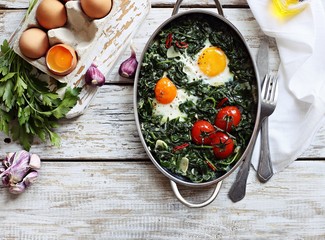 Eggs baked with spinach and tomatoes in serving pan overhead rustic wooden table. Overhead view.