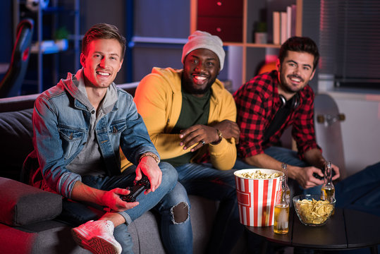 Best friends. Portrait of cheerful stylish young guy is resting on couch with joystick while looking at camera with joy. Positive men are sitting in background and expressing gladness