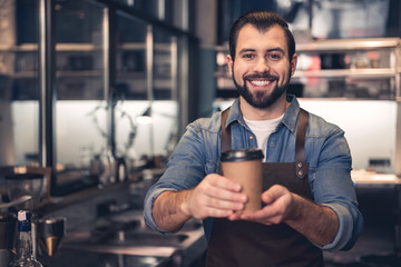 Portrait of beaming unshaven barista keeping mug of delicious coffee in hand while locating in confectionary shop. Job concept - obrazy, fototapety, plakaty