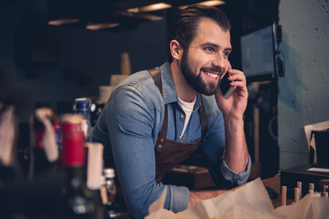 Portrait of outgoing bearded barista speaking on mobile while leaning on bar counter in cafe. Break...