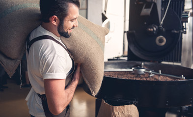 Side view cheerful unshaven professional coffee roaster worker holding bag in hands while going near technical equipment at factory. Industry concept